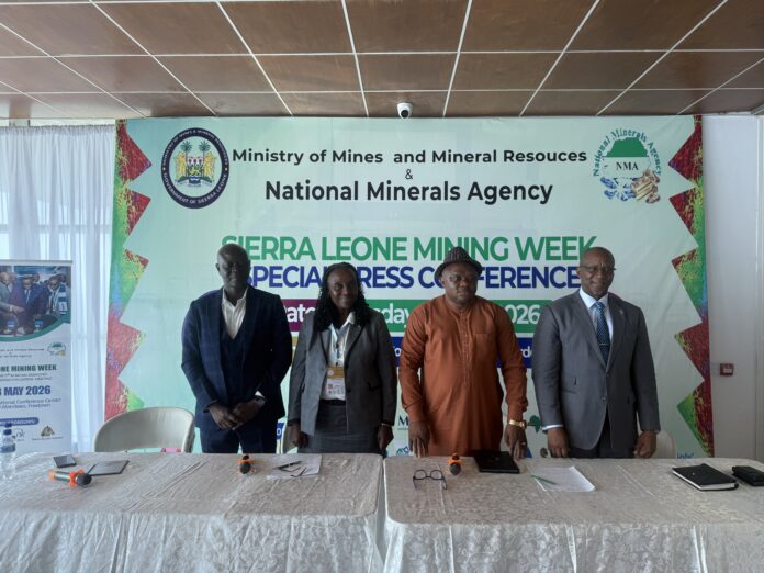 Four professionally dressed individuals stand behind a long, white-clothed table in a conference setting, with a large banner reading 'Ministry of Mines and Mineral Resources & National Minerals Agency' in the background and microphones on the table in front of them.