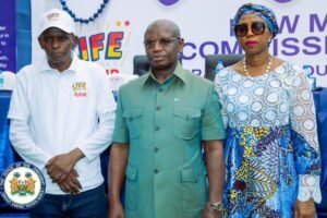 Three adults pose for a photo at a formal event: left man in a white Life Flour polo, middle man in a green button-up, right woman in a blue patterned dress with headwrap and sunglasses.