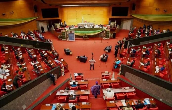 Large legislative chamber with curved red seats, attendees seated at desks facing a central stage and podiums atop a long table.