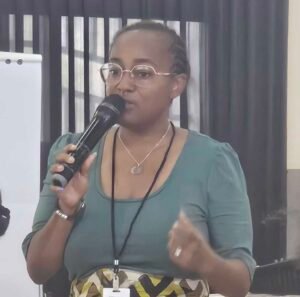 Woman with glasses speaks into a handheld microphone during a presentation, standing near a whiteboard.
