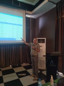 Man giving a presentation: pointing to a projected slide in a conference room, holding a microphone beside a laptop and podium.