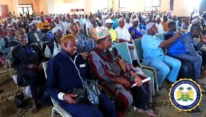 Large crowd seated in a hall for an official event; front-row men wear traditional attire and hats