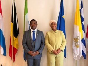 Two formally dressed individuals stand side by side in front of a row of flags, posing for a photo in a formal setting.