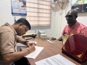Two men in an office: one signs a document at a wooden desk while the other, wearing a red patterned shirt and sunglasses, watches nearby.