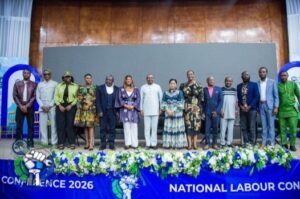 Group of diverse men and women standing on a stage for a conference photo; banner reads 'National Labour Conference 2026' with blue floral decorations across the front.