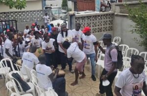 Crowd of people in white shirts at an outdoor community event; some hold posters, others drum and cheer amid plastic chairs and a low wall.