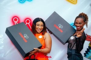 Two smiling women at a brand event pose with oversized black prize boxes featuring a logo on each box.