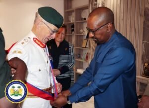 Man in blue traditional attire ties a red ceremonial sash around a uniformed officer's waist during a formal indoor ceremony; badge seal visible in corner.