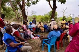 A diverse group of people sitting in a circle under trees, holding a community meeting outdoors.