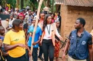 A diverse group of community members walk through a village as a woman in an orange safety vest leads with a tall bamboo pole in hand.