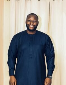 Smiling man in a navy traditional tunic standing in front of cream curtains.