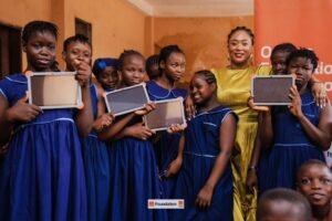 Group of girls in blue uniforms holding tablet devices with a smiling woman in yellow at a school event.