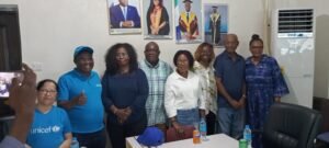 Group of eight adults posing in a meeting room; UNICEF staff in blue shirts, posters on the wall behind, water bottles on the table
