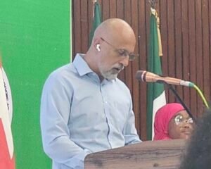 Man with gray beard and glasses speaks at a wooden podium, wearing a light blue shirt and wireless earbud, into a microphone. Nearby is a woman in a pink headscarf.