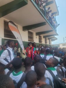 Crowd of students and staff lining a school building’s exterior, watching a long orange-and-white banner or float held between them.