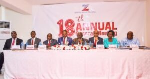 Group of eight professionals seated behind a long pink table on a stage at an annual conference, banner behind them reads '18th Annual' and shows a corporate logo.