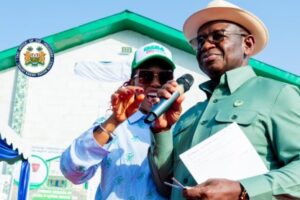 Two officials at an outdoor ceremony speak into a microphone in front of a new building, with a government seal visible on the wall.