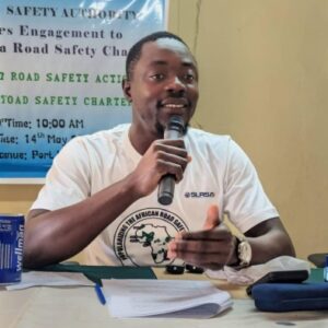Man speaking into a microphone at a road safety event, seated at a table with papers and a banner behind him.