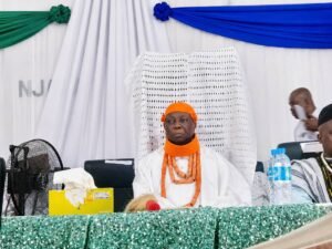 Man in white traditional attire with an orange head wrap and bead necklace sits at a ceremonial dais.