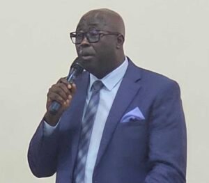 | The Calabash Newspaper Man in a navy suit with a light blue pocket square, speaking into a handheld microphone at a formal event.