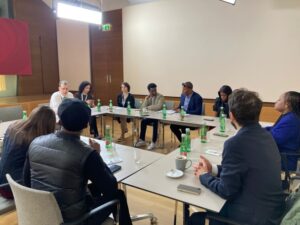 Roundtable meeting with diverse professionals seated around a U-shaped table in a conference room, bottles of water and cups on the table, engaging discussion underway.