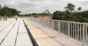 Construction workers walk along a partially completed bridge deck with metal railing on the right, over a river with trees in the distance.