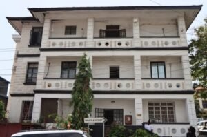 | The Calabash Newspaper Three-story white residential building with balconies on each floor, circular decorative cutouts on the façade, a tall tree in front, and a street sign reading Walpole Street near the entrance.