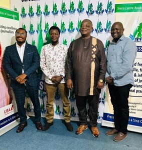 Four men stand side by side posing for a photo at a Sierra Leone journalists event, with banners in the background.