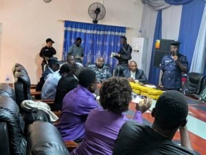 Group of people in a formal meeting around a conference table with yellow packages in the center, a police officer speaking into a microphone at the right side.