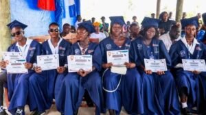 Group of graduates in blue gowns seated outdoors, each holding a certificate from a graduation ceremony.
