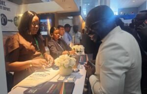 Smiling man in a white suit talking to a booth staffer at a conference expo, with brochures and white flower centerpiece on the table