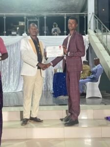 Two men in suits shake hands on a stage as one receives a certificate at an awards ceremony, with a draped backdrop and seated guests in the background.