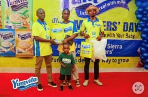 Group photo at a Yumvita infant cereal promotion: four adults in yellow shirts and a young child pose on a red carpet with product boxes and large banner in the background to promote the brand.