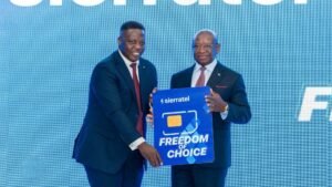 Two men in dark suits on a blue stage hold a large blue poster that reads 'FREEDOM OF CHOICE' and features a SIM card graphic and Sierra Tel branding.