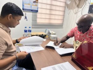 Two men sit across a desk in an office, one flipping through documents while the other signs papers on the table in front of them.