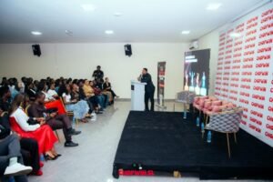 Speaker at podium addressing a seated audience in a conference room with a stage and promotional banners outside the room viewledpresents scene