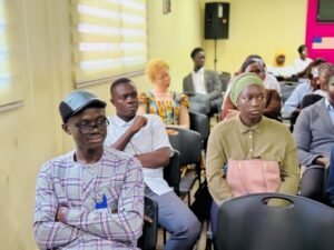 Diverse group of adults seated in a classroom or meeting room, listening attentively to a presentation.