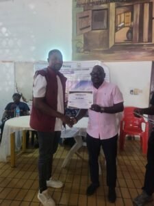 Two men stand indoors as one hands a certificate to the other during a small award ceremony, with a table and mural in the background.