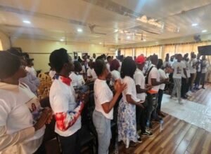| The Calabash Newspaper People standing in rows and clapping inside a large indoor hall, many wearing white shirts and facing a stage.