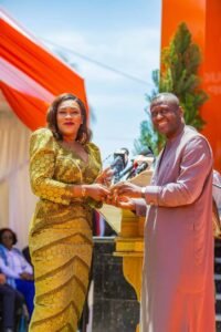 Woman in a gold sequined dress presents an award to a man in a light purple traditional robe at an outdoor ceremony with orange tents in the background.