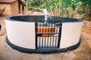 Circular concrete water well with a black fence gate; orange pump mechanism inside, taped off with yellow and blue caution tape in a rural yard.