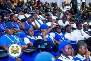 Crowd of people seated at an event, many in blue uniforms and berets; government seal visible in the lower left.