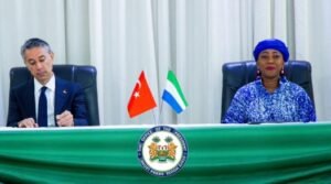 Two officials sit at a green-cloth table; the man on the left signs a document while the woman on the right sits attentively. Between them are two small flags (one red with a white star and crescent, the other green/white/blue) and a circular official seal on the front of the table.