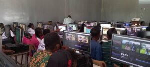 A crowded computer lab with students at desks editing videos on large monitors in a classroom setting.