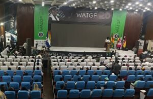Conference hall with a stage, WAIGF banners, and flags; audience seated in rows facing the stage.