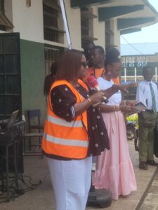 Group of people outdoors around a table; a woman in an orange safety vest hands a document or item to a man in a white shirt while others watch, some in formal attire including a pink dress, in a campus or building courtyard setting.
