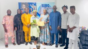 Group of nine people in colorful traditional attire posing in a studio with SLBC banners in the background; a certificate is being handed to a woman in a green headscarf in the center of the group.