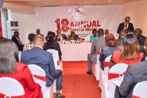 Panel of speakers sits behind a long table on a stage, addressing an audience at an 18th Annual General Meeting.