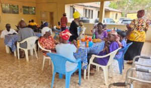 People dining together at a covered outdoor space, colorful clothing, blue-check tablecloths, and plastic chairs share a communal meal.