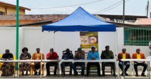 Row of people seated behind a long white table under a blue canopy at an outdoor community event; banner with NP logo in the background.